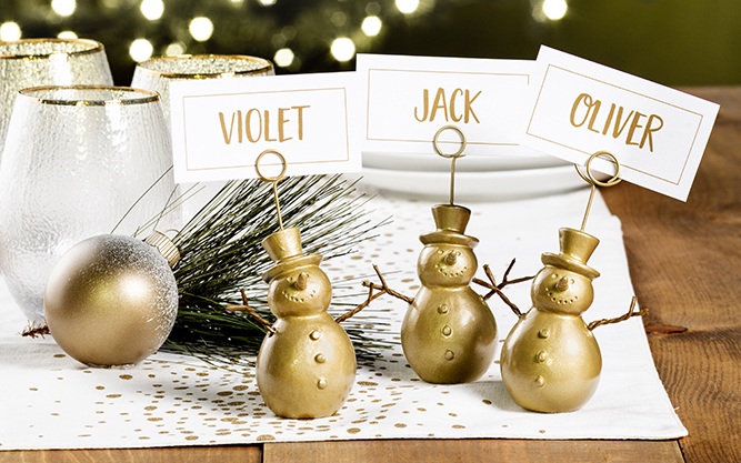 Three gold snowman place card holders with cards reading Violet, Jack and Oliver on a table with a Christmas tree in the background.