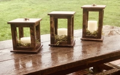 Three wooden lanterns holding candles on top of an outdoor wooden table