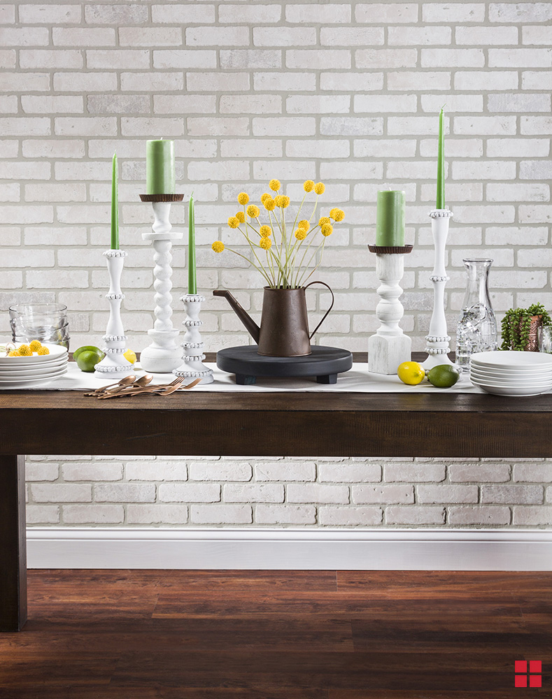 Table with white plates, white candlesticks with green candles and a centerpiece of yellow flowers in a copper pitcher sitting on a black trivet.