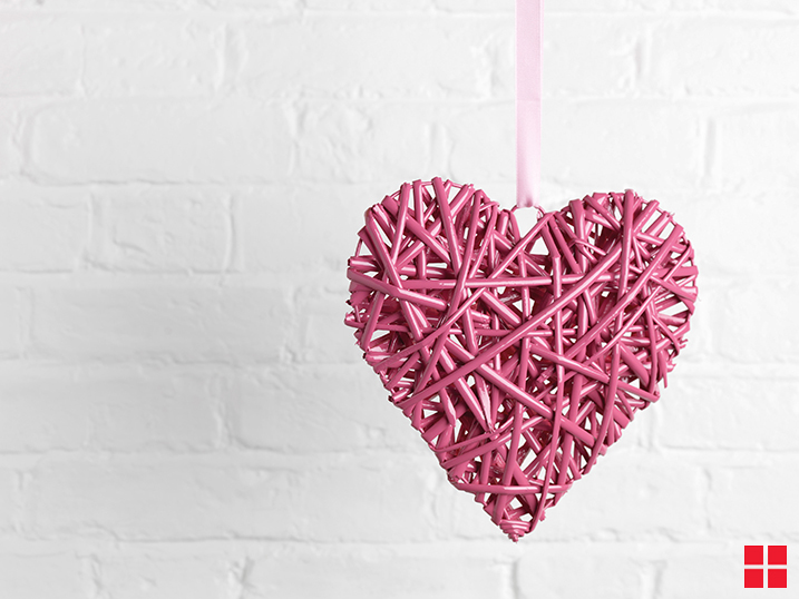 A pink wicker heart hanging from a pink ribbon in front of a white brick wall.